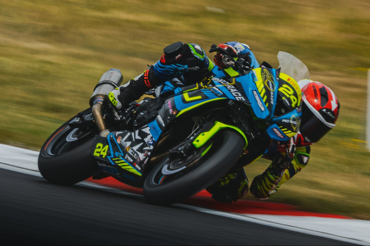 A motorcycle racer leans sharply into a turn on a racetrack, wearing a blue, black, and neon green racing suit with a matching helmet. The blue and black racing bike is angled low over the track surface, with a blurred grassy infield and track barriers in the background.